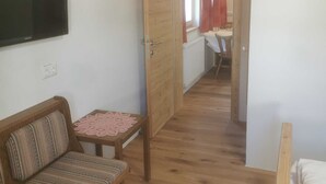 A small, cozy room with a wooden floor, a sofa, a table with a lace tablecloth, a television on the wall, and a door leading to another room.