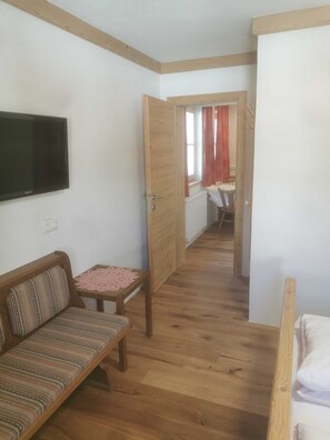 A small, cozy room with a wooden floor, a sofa, a table with a lace tablecloth, a television on the wall, and a door leading to another room. - Ferienwohnung 5 - Gästehaus Obererlacher (Obertilliach)