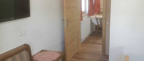 Une petite pièce chaleureuse, avec un plancher en bois, un canapé, une table recouverte d’une nappe en dentelle, un téléviseur accroché au mur et une porte donnant sur une autre pièce.