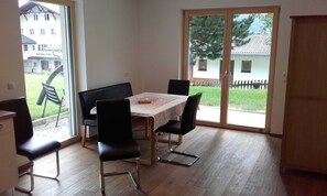 Un coin repas avec une table et des chaises, une vue sur un bâtiment portant l’inscription « GEMEINDE » à travers une porte vitrée, et un plancher en bois.