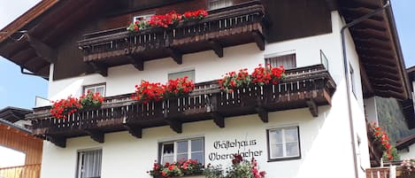 Un immeuble à plusieurs étages doté de balcons ornés de fleurs rouges, une enseigne indiquant « Balkhaus Obererwacher », et une structure en rondins de bois au premier plan.