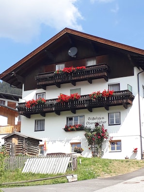 Un edificio de dos plantas con balcones decorados con flores rojas, un letrero que dice 'Käfighaus Oberercher' y una estructura de cabaña de troncos al frente.