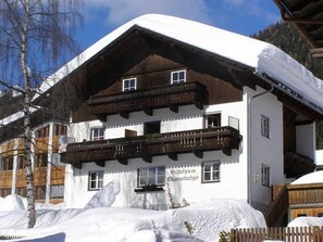 Ein traditionelles Chalet mit Holzbalkon, einem Schild, das es als Pension kennzeichnet, und verschneiter Umgebung.