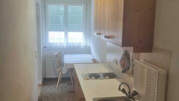 A compact kitchen with a white countertop, a sink, wooden cabinets, a radiator, and a window with blinds.