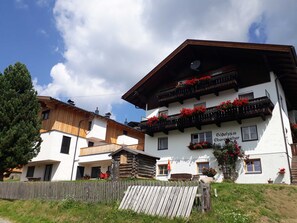 Il s’agit d’un bâtiment de style chalet traditionnel, avec une façade en bois, des balcons ornés de fleurs rouges et une enseigne indiquant qu’il s’agit d’une maison d’hôtes.