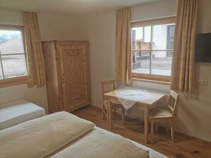Una habitación con un armario de madera, una mesa con sillas, una cama y un ventanal con cortinas.