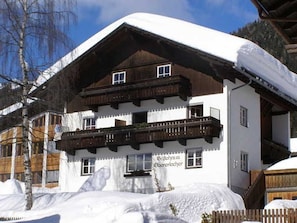 A traditional chalet with a wooden balcony, a sign indicating it's a guesthouse, and snow-covered surroundings.