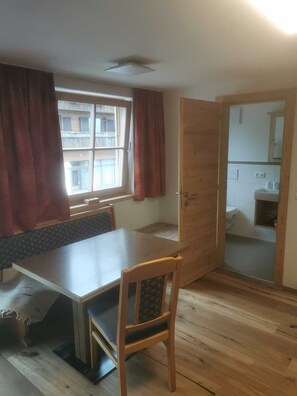 Een kamer met een houten eettafel, een enkele stoel, een raam met gordijnen en uitzicht op een gebouw.