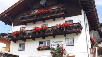 Ein mehrgeschossiges Gebäude mit Balkonen, die mit roten Blumen geschmückt sind, eine Schrifttafel mit der Aufschrift 'Balkhaus Oberwacher' und eine Blockhütte im Vordergrund.