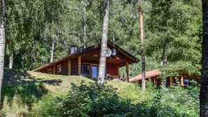 Une cabane en bois avec un toit en pente, de grandes fenêtres et des antennes paraboliques, entourée d’une forêt dense.