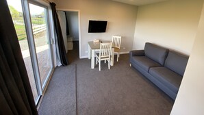 Ein Zimmer mit grauem Sofa, einem Tisch mit Stühlen, einem an der Wand montierten Fernseher und einer Glastür, die nach draußen führt.