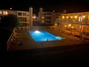 A swimming pool area with a fenced deck, a covered seating area, and a building with multiple windows and balconies.