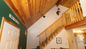 Eine Treppe mit Holzgeländern führt zu einer zweiten Etage, eine grüne Wand mit dem Schild 'Family Friends Traditions' und ein braunes Sofa im Vordergrund.