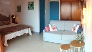A hotel room with a bed, a sofa, two small tables, a wall-mounted air conditioner, and a door.