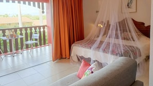 A bedroom with a bed under a mosquito net, a balcony with a table and chairs, and a view of greenery outside.