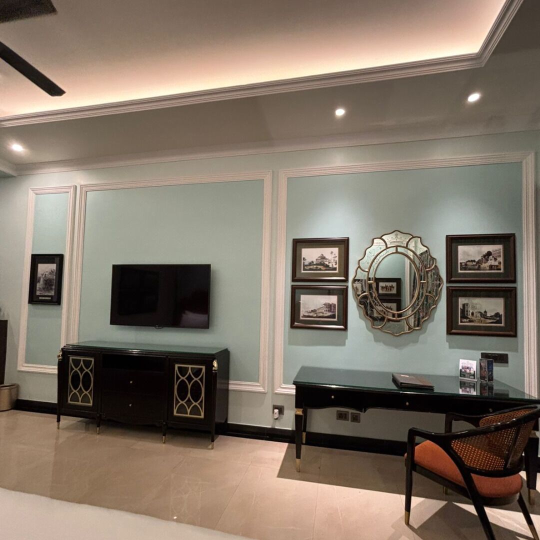 A room with a flat-screen TV mounted on the wall, a black console table, a glass-top desk, and a wicker chair.