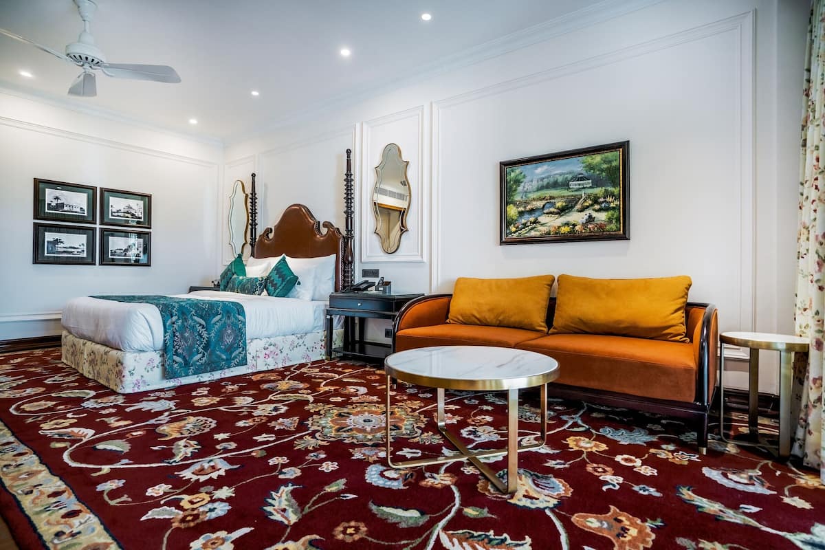 A room with a bed, a sofa, a round coffee table, a ceiling fan, framed pictures, and a painting.