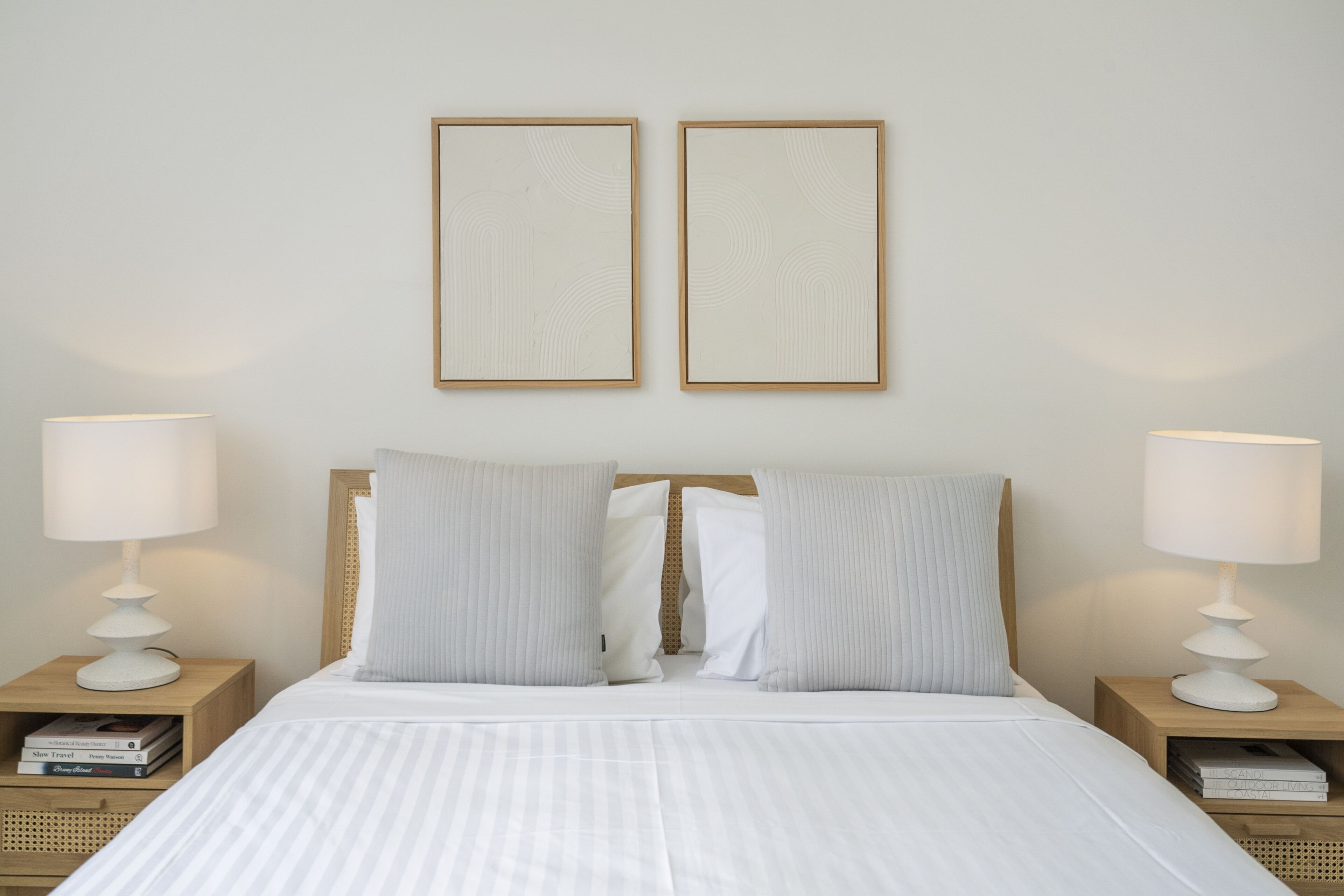 Una cama bien tendida con ropa blanca, flanqueada por dos mesitas de noche con lámparas y libros.
