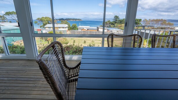 A wooden deck with wicker chairs and a table, overlooking a coastal area with houses and a view of the sea. - Blue Cottage Bruny Island (Alonnah)