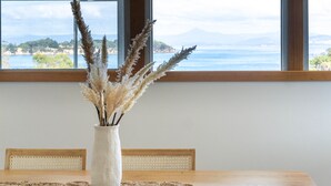 A dining table with a vase of pampas grass, a woven placemat, and a view of a coastal landscape through a window. - Blue Cottage Bruny Island (Alonnah)