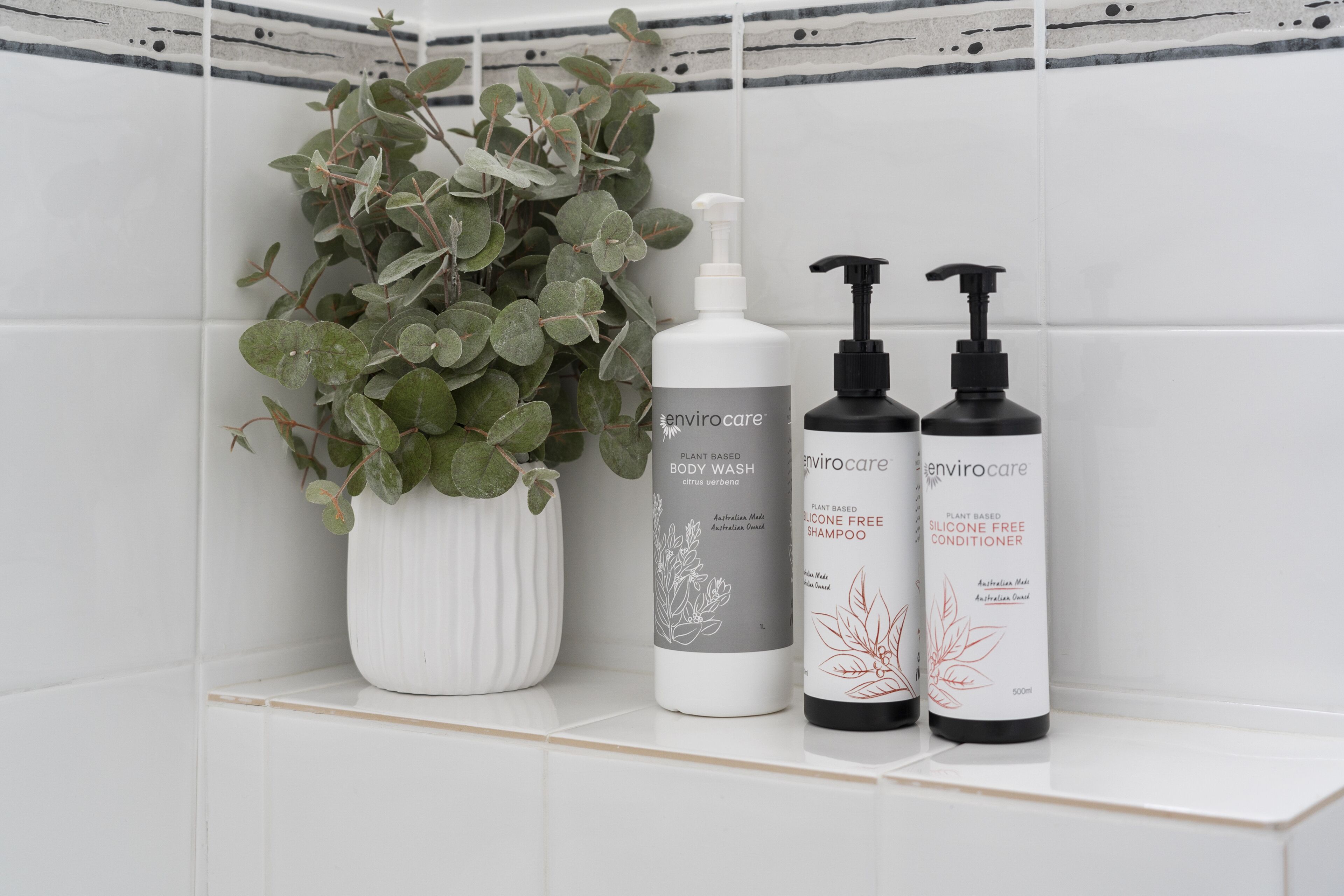 A bathroom shelf with a white vase containing green foliage, a body wash bottle, and two conditioner bottles.