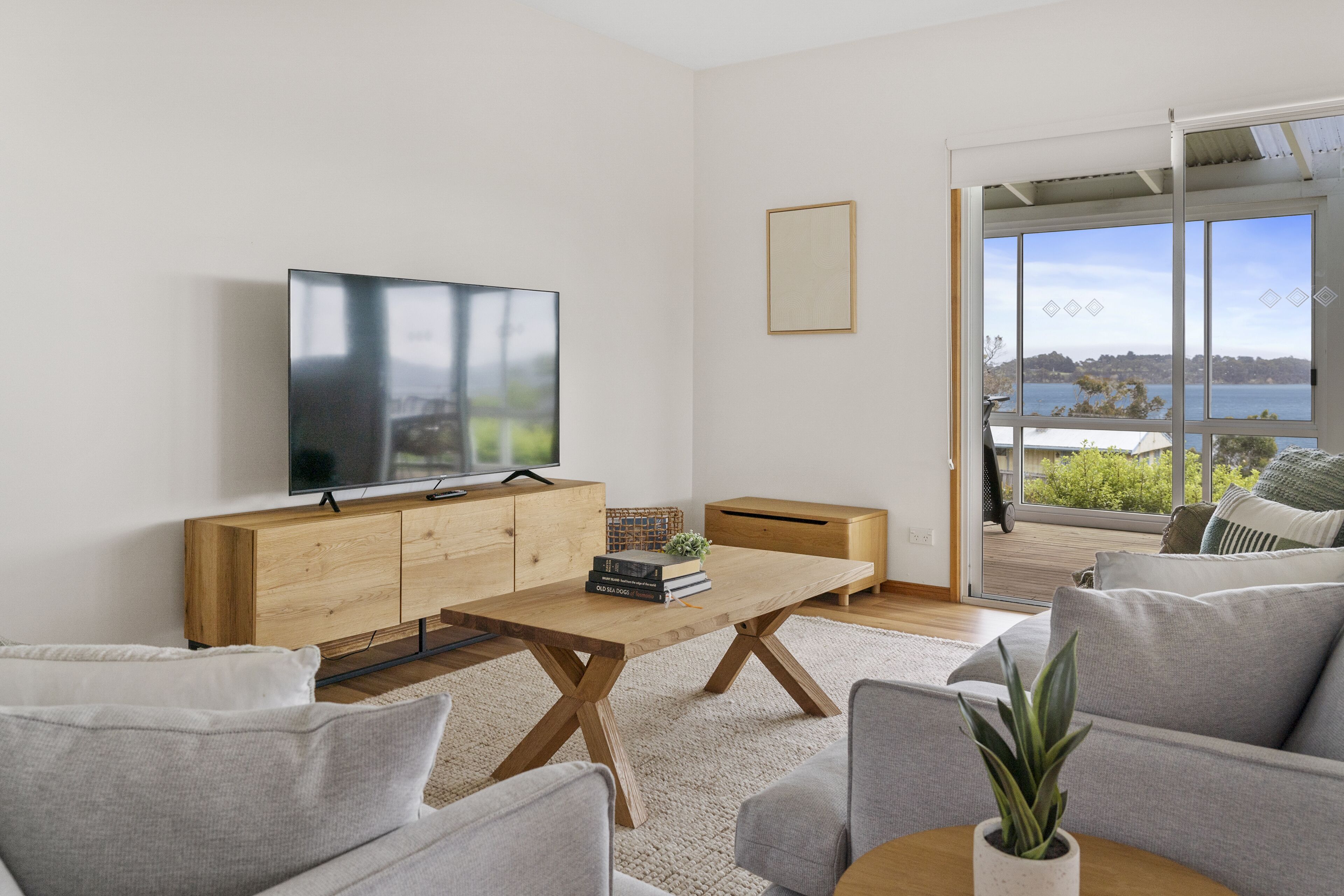 A modern living room with a wooden coffee table, grey sofas, a flat-screen TV, and a large window offering a view of a body of water.