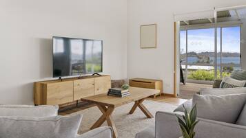 A modern living room with a wooden coffee table, grey sofas, a flat-screen TV, and a large window offering a view of a body of water.