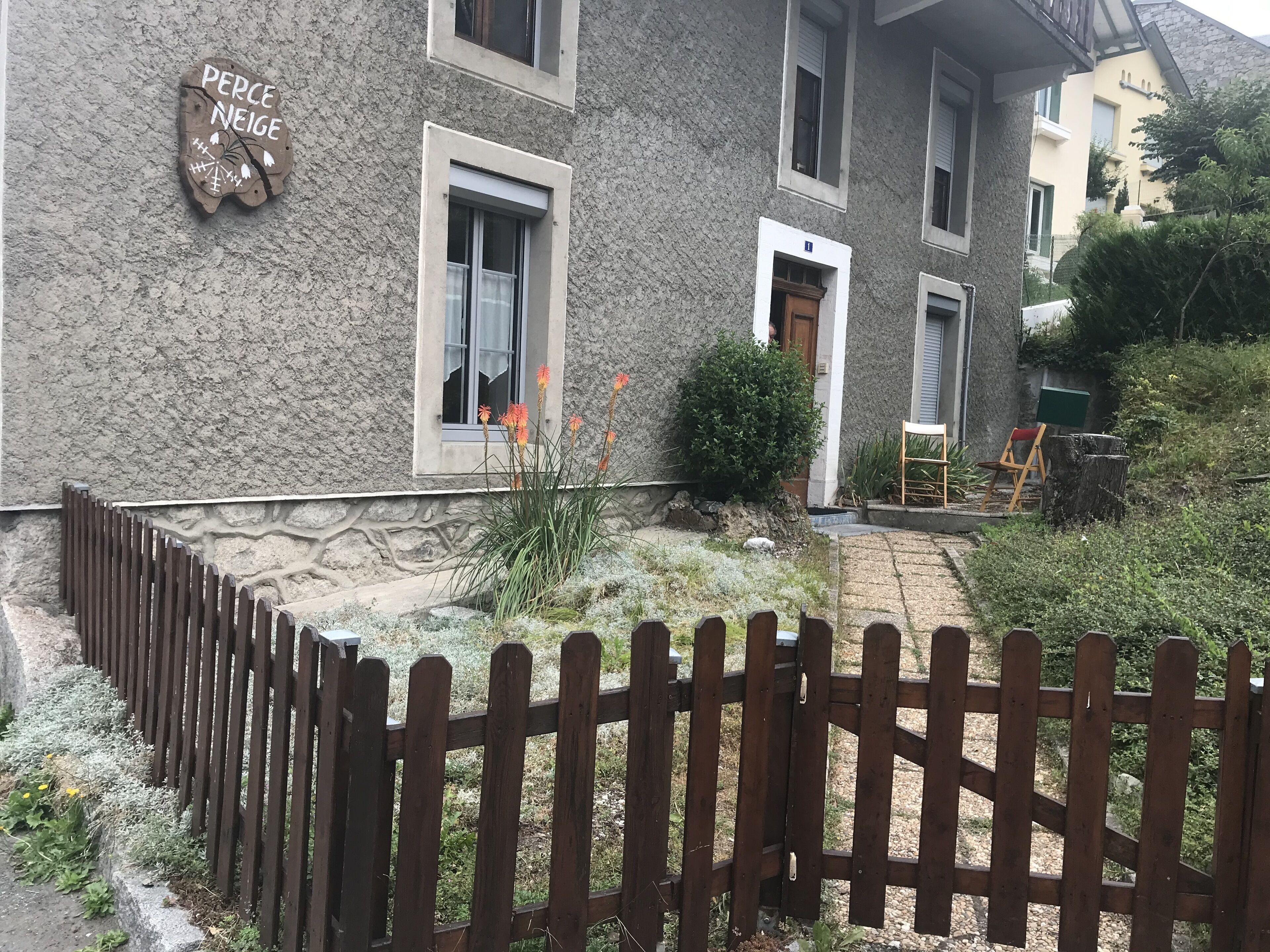 Un bâtiment en pierre avec une clôture en bois, un petit jardin avec des fleurs oranges et une porte en bois.