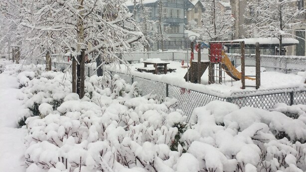 A snow-covered park with playground equipment, a fence, and buildings in the background.