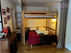 A compact room with a bunk bed, wooden walls, a small desk with a red kettle, and a shelf with framed pictures.