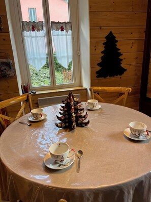 A round wooden table with a tablecloth, surrounded by four chairs. There are four cups and saucers with reindeer designs, a spoon, and a decorative tree-shaped object on the table.