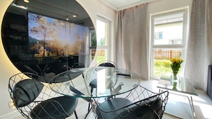 Ein moderner Essbereich mit einem Glastisch und Drahtgestellstühlen, einem großen Flachbildfernseher und Blick auf einen Garten durch ein Fenster.