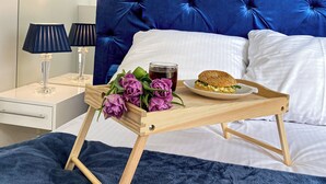 Ein Bett mit blauem Kopfteil, weißen Kissen, einem Holztablett mit einem Burger und Blumen sowie einem Nachttisch mit Lampen.