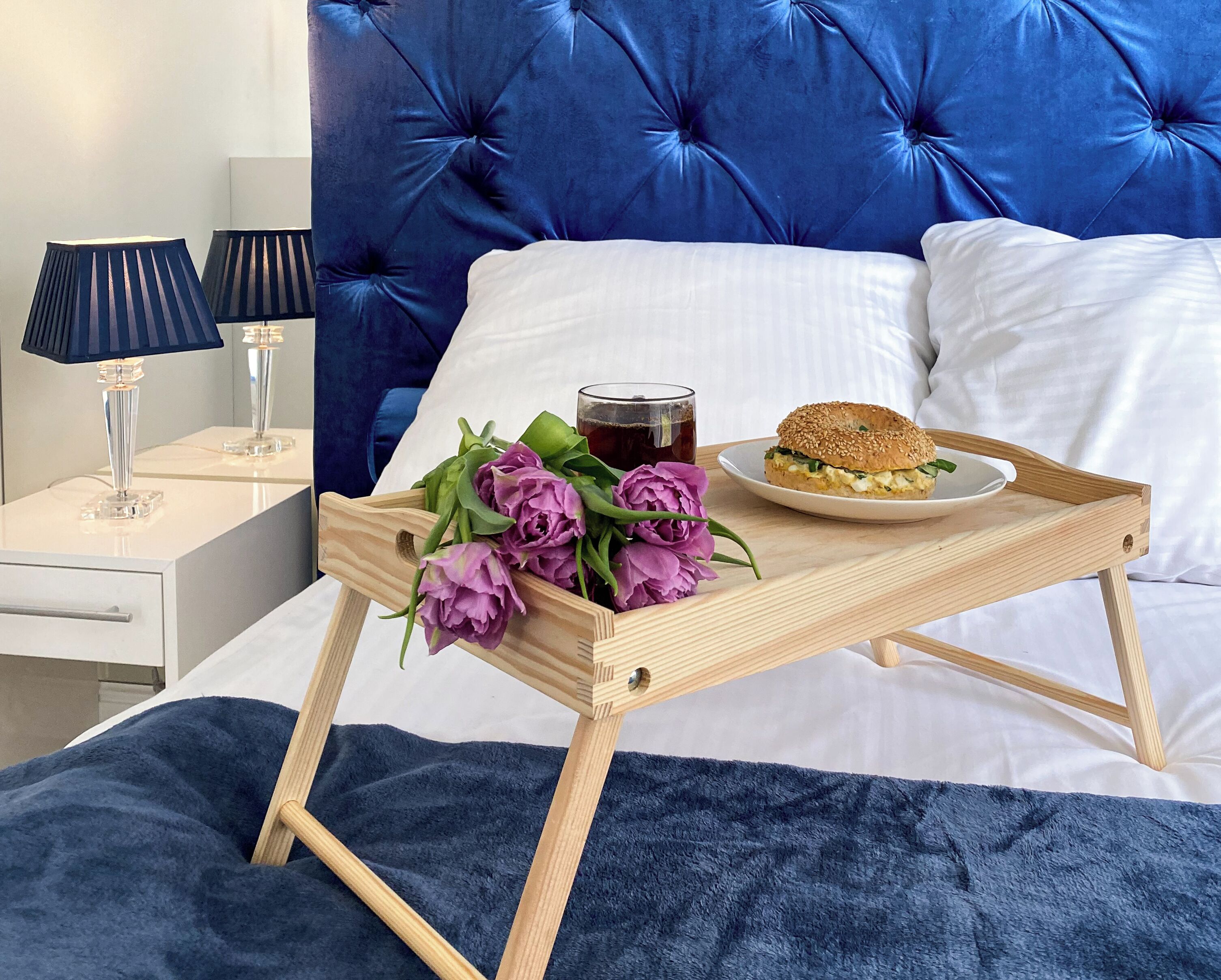 A neatly made bed with a blue headboard, a bedside table with a lamp, and a tray with flowers, a drink, and a sandwich.