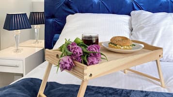 A neatly made bed with a blue headboard, a bedside table with a lamp, and a tray with flowers, a drink, and a sandwich.