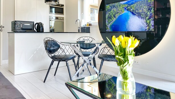 Eine moderne Küche mit weißen Schränken, einem eingebauten Backofen und einer Mikrowelle. Es gibt einen Essbereich mit einem Glastisch und schwarzen Stuhlgestellen. Ein großer Flachbildfernseher ist an der Wand montiert und zeigt eine idyllische Landschaft.