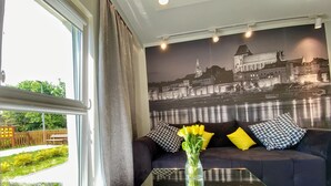 Ein Wohnzimmer mit großem Fenster, einem Glastisch, einer dunklen Couch mit gemusterten Kissen und einem Wandgemälde mit Stadtansicht.