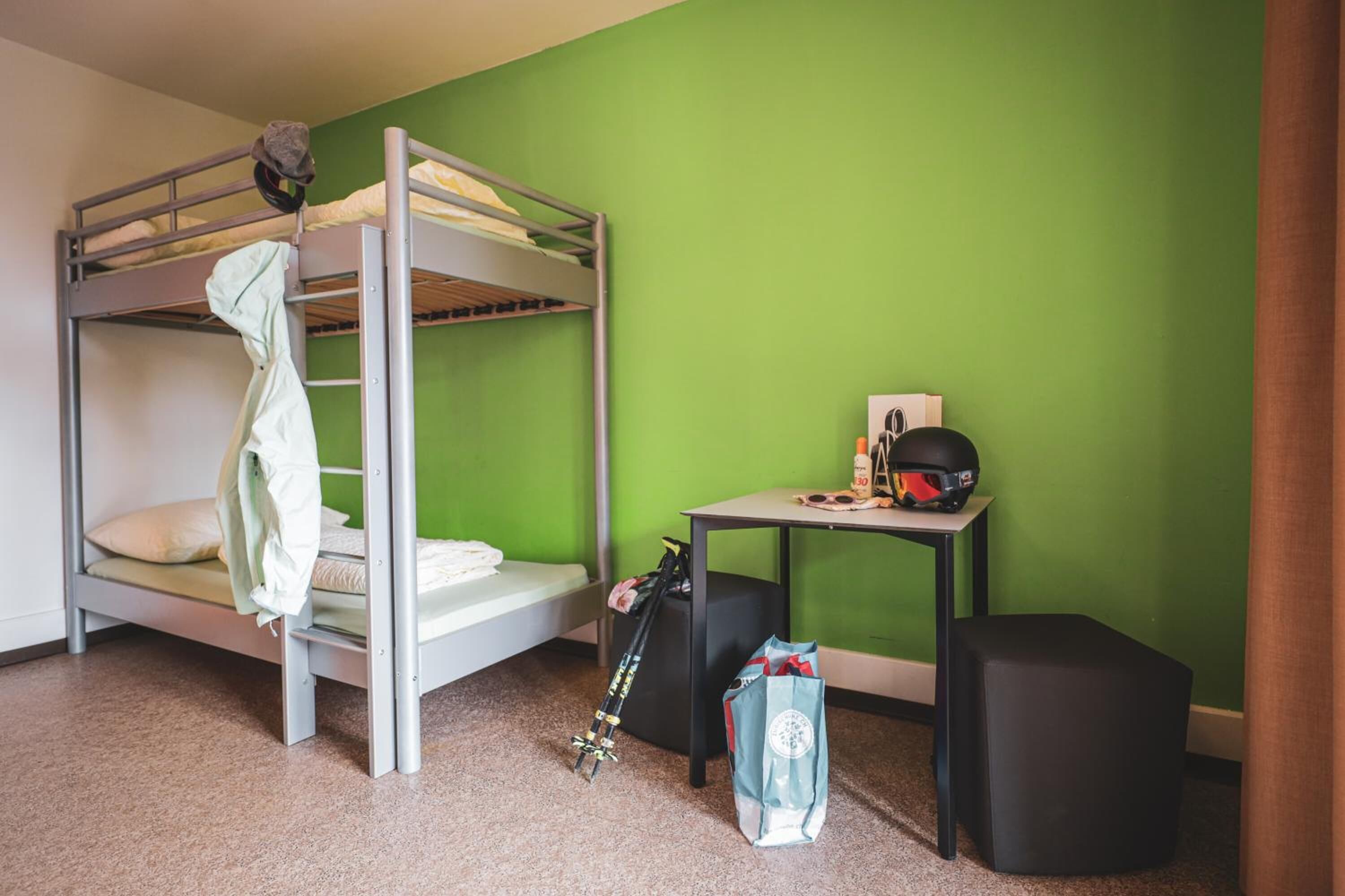 A bunk bed room with a green wall, a small table with a helmet and sunglasses, and a bag on the floor.