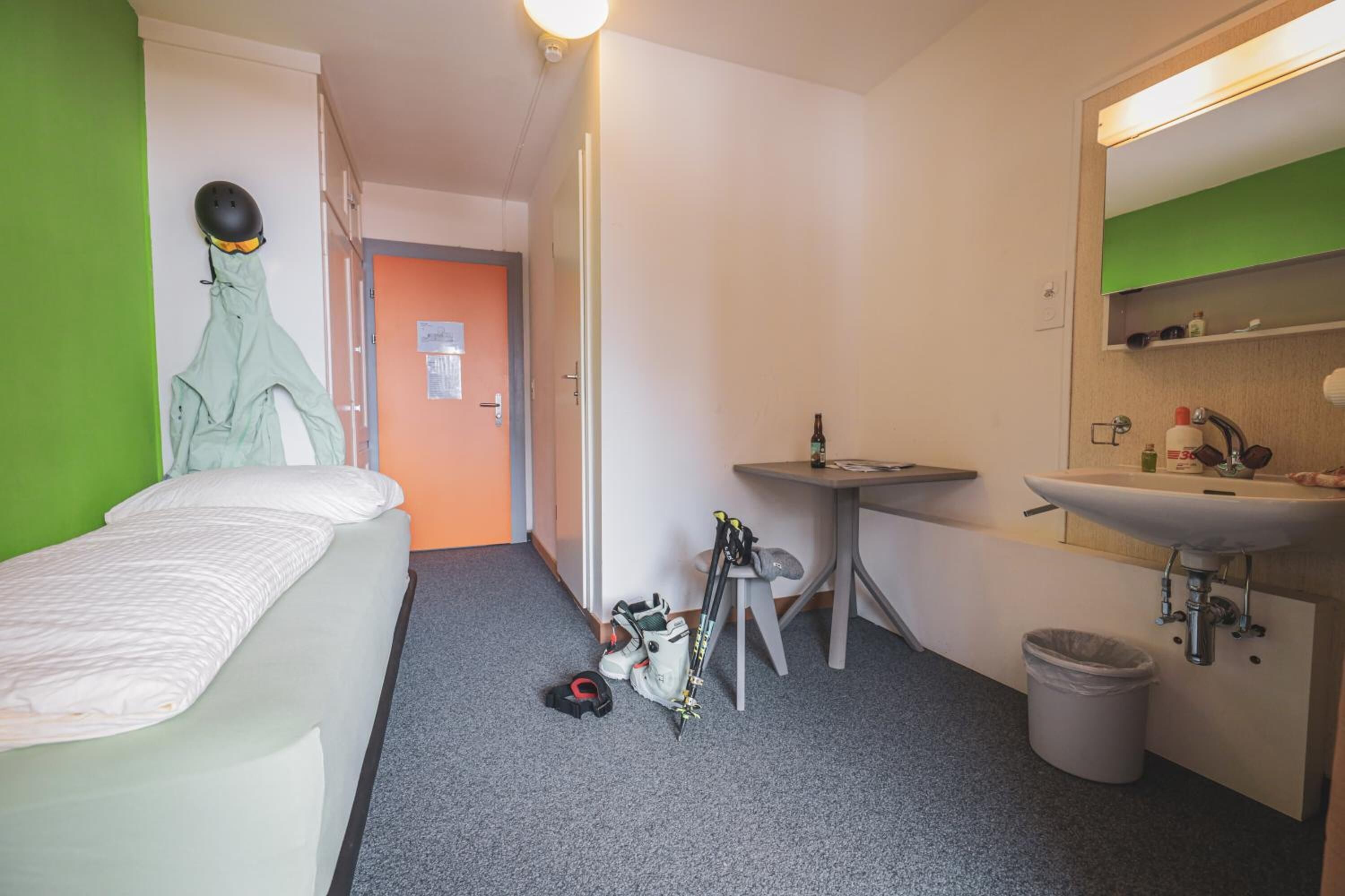Et lille hotelværelse med seng, skrivebord, stol, håndvask og spejl.