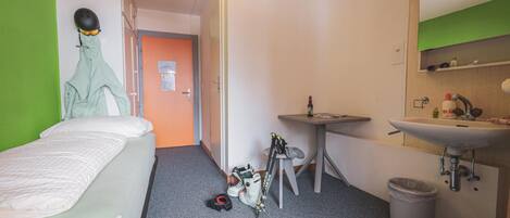 Ein kleines Hotelzimmer mit Bett, Schreibtisch, Stuhl, Waschbecken und Spiegel.
