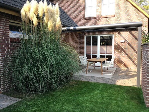 Une maison en briques avec un patio, une table et des chaises, et de hautes herbes.