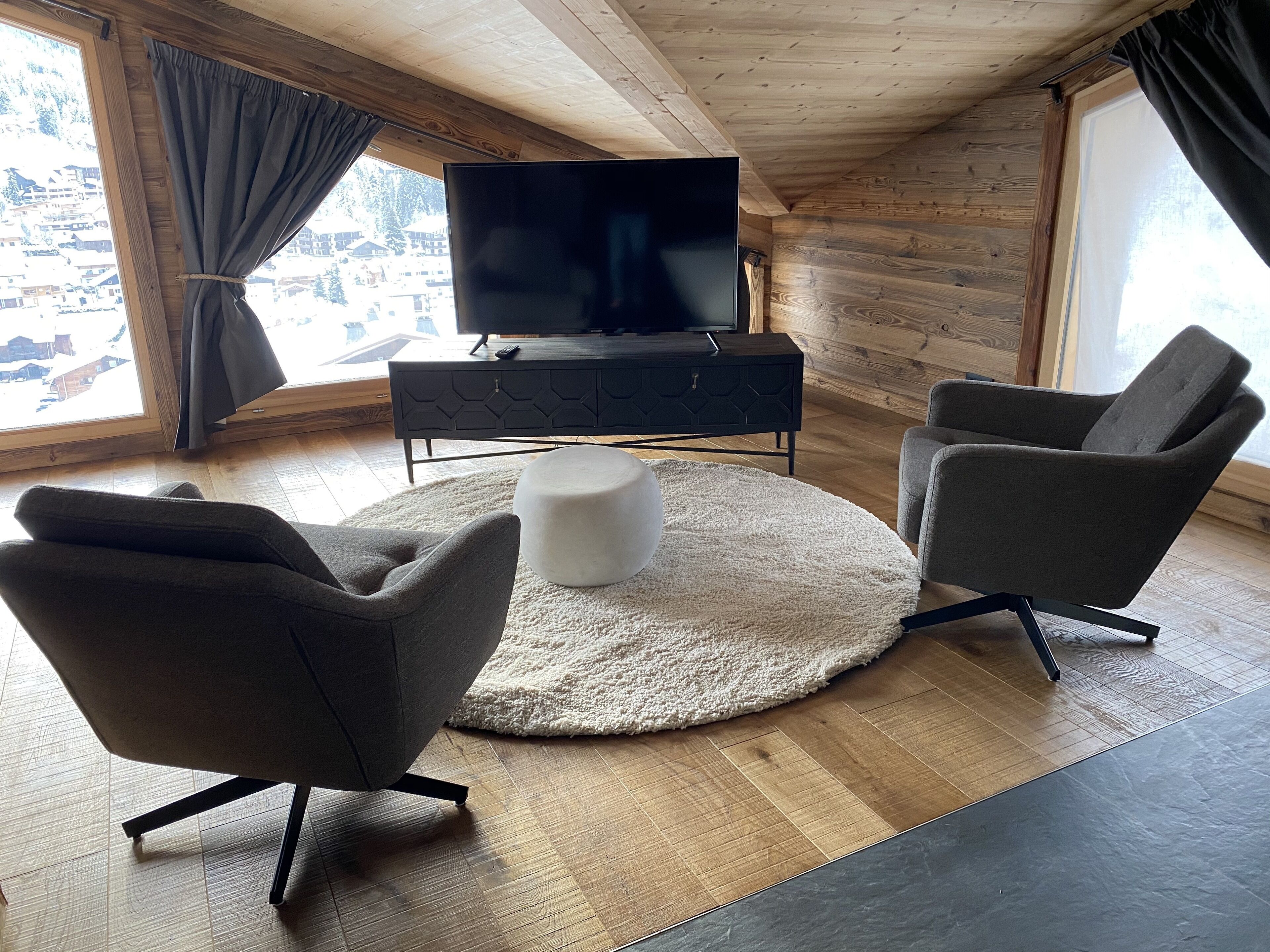 A modern living room with a television, two armchairs, a round ottoman, and a wooden floor.