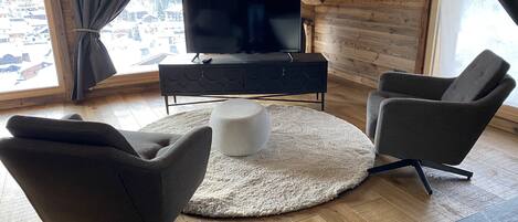 A modern living room with a television, two armchairs, a round ottoman, and a wooden floor.