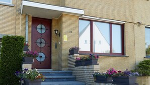 Un bâtiment à deux étages avec une porte rouge, de nombreuses fenêtres et des marches menant à l’entrée. Des jardinières sont placées sur les marches et une boîte aux lettres se trouve sur le côté droit.