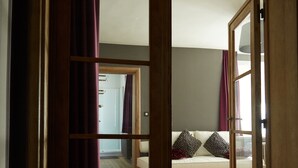 Une porte en bois ouverte donne sur une pièce où se trouvent un lit, un miroir et d’où l’on peut voir une autre pièce.