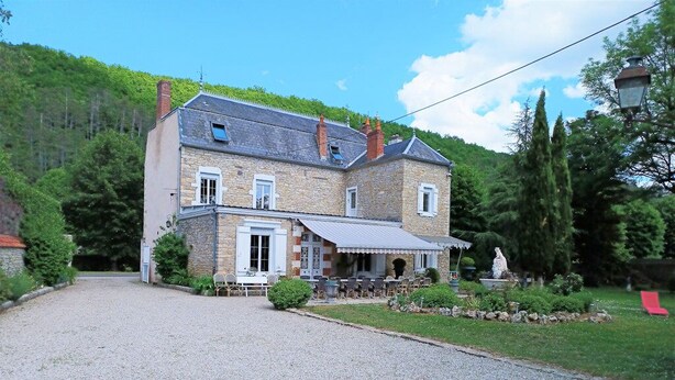 Une maison en pierre à deux étages, avec une allée gravillonnée, un espace extérieur aménagé pour s’asseoir et une statue dans le jardin.