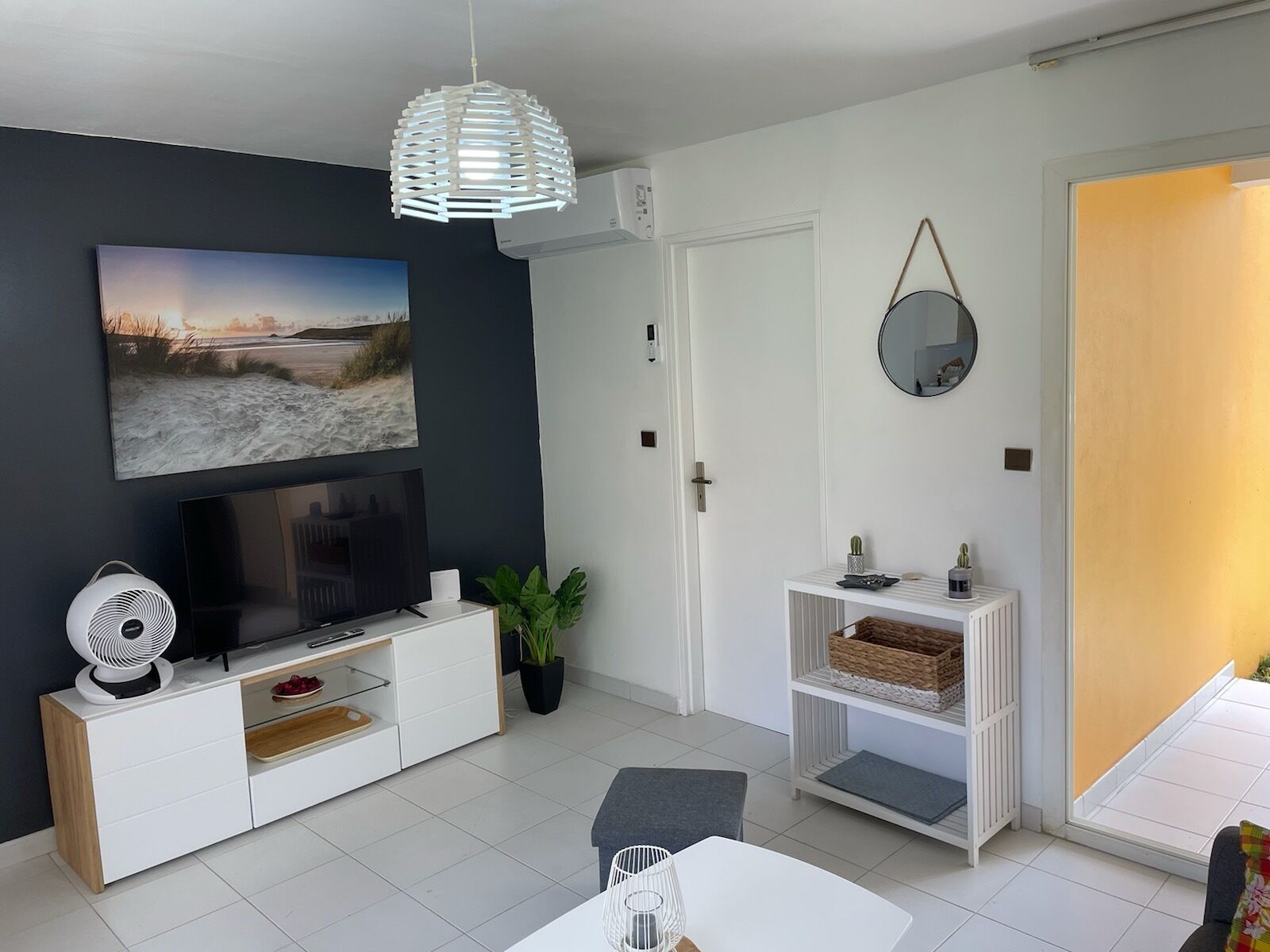 Una sala moderna con televisor de pantalla plana, ventilador de techo, planta en maceta y comedor con mesa y sillas blancas.
