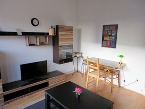 Un salon moderne avec un téléviseur à écran plat, des meubles en bois, une table à manger avec des chaises, une petite table ornée d’un vase de fleurs et une horloge au mur.