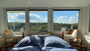 A bedroom with a bed centered between two windows, offering a view of a green landscape. - Exceptional vacation home in a fantastic location above Rheinfels Castle, Loreley (Sankt Goar)