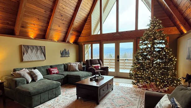 Una sala de estar acogedora con techo de madera, un ventanal grande, un sofá verde, una mesa de centro y un árbol de Navidad.
